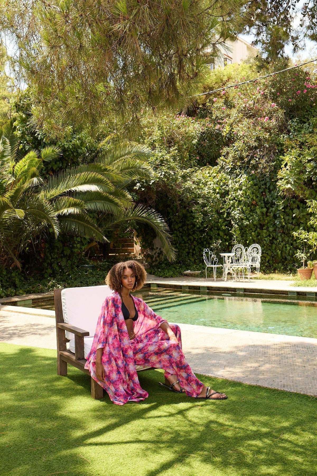 Woman in a colorful dress sitting on a bench by a pool with greenery in the background