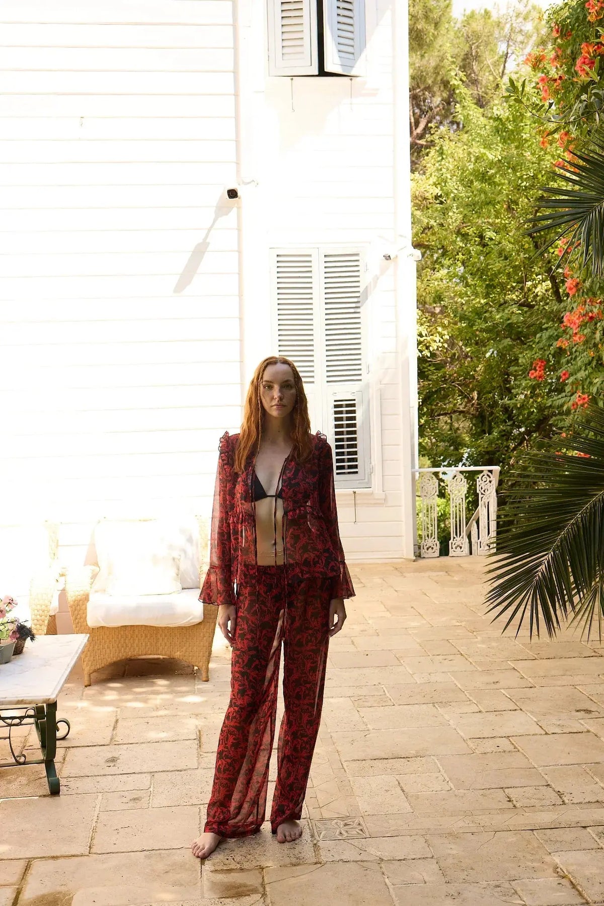 Woman in a red outfit standing on a patio with greenery and a white building in the background