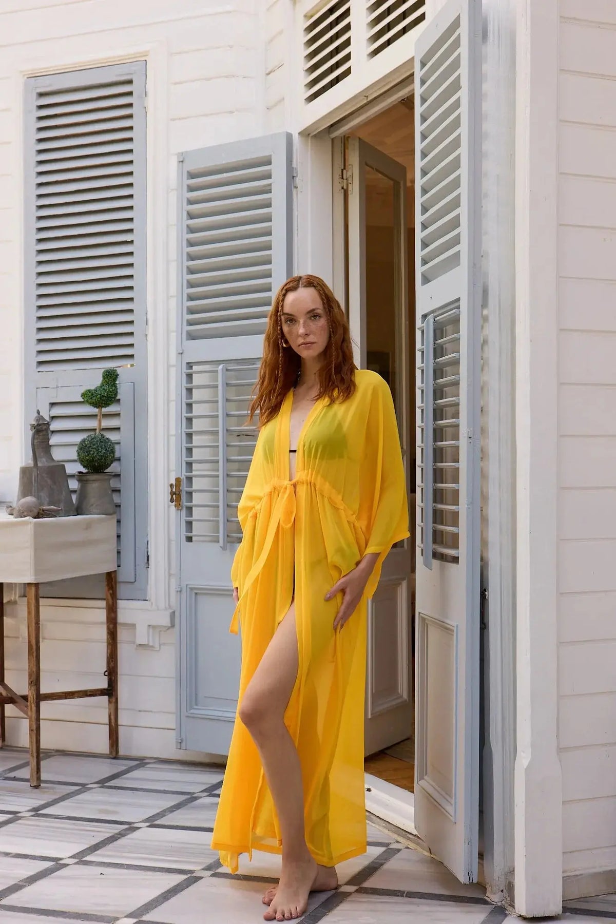Woman in a yellow dress standing in front of a white building with blue shutters.
