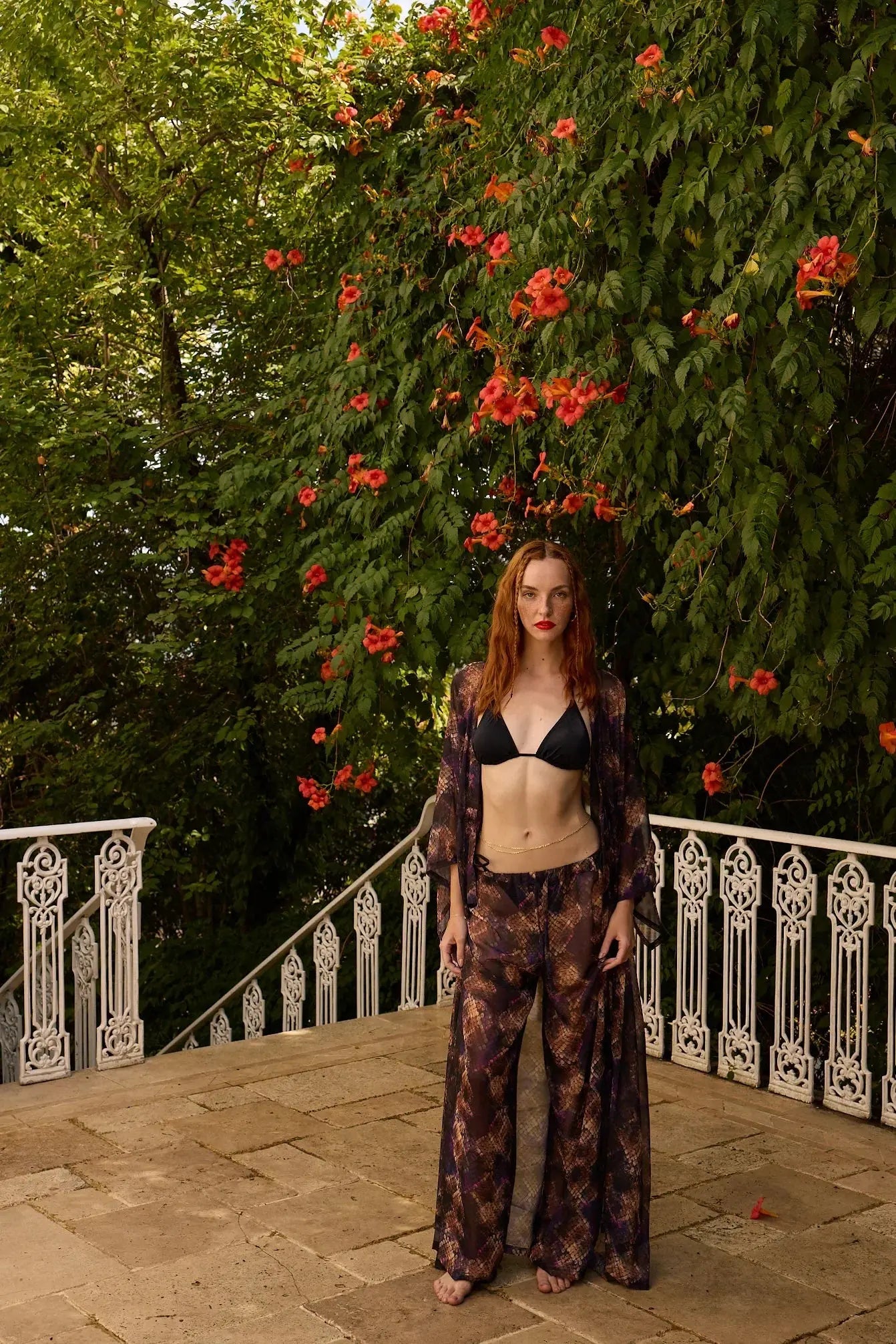 Woman standing on a stone patio with a lush green wall and red flowers in the background