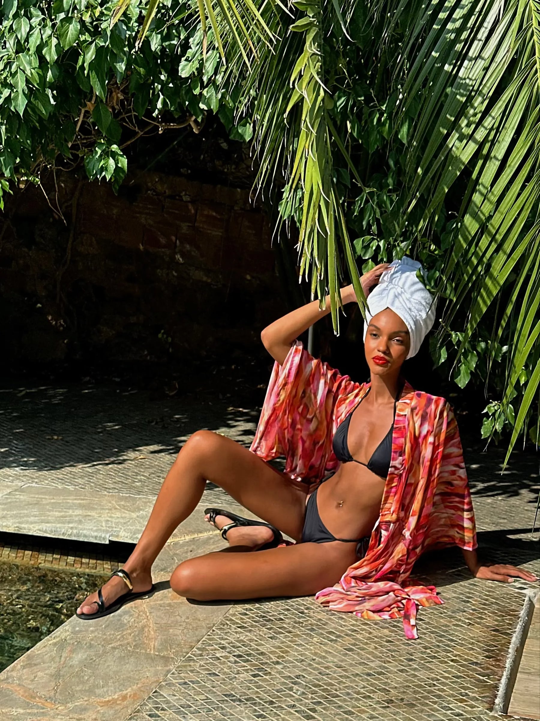 Woman in a bikini and colorful robe sitting by a pool with tropical plants in the background