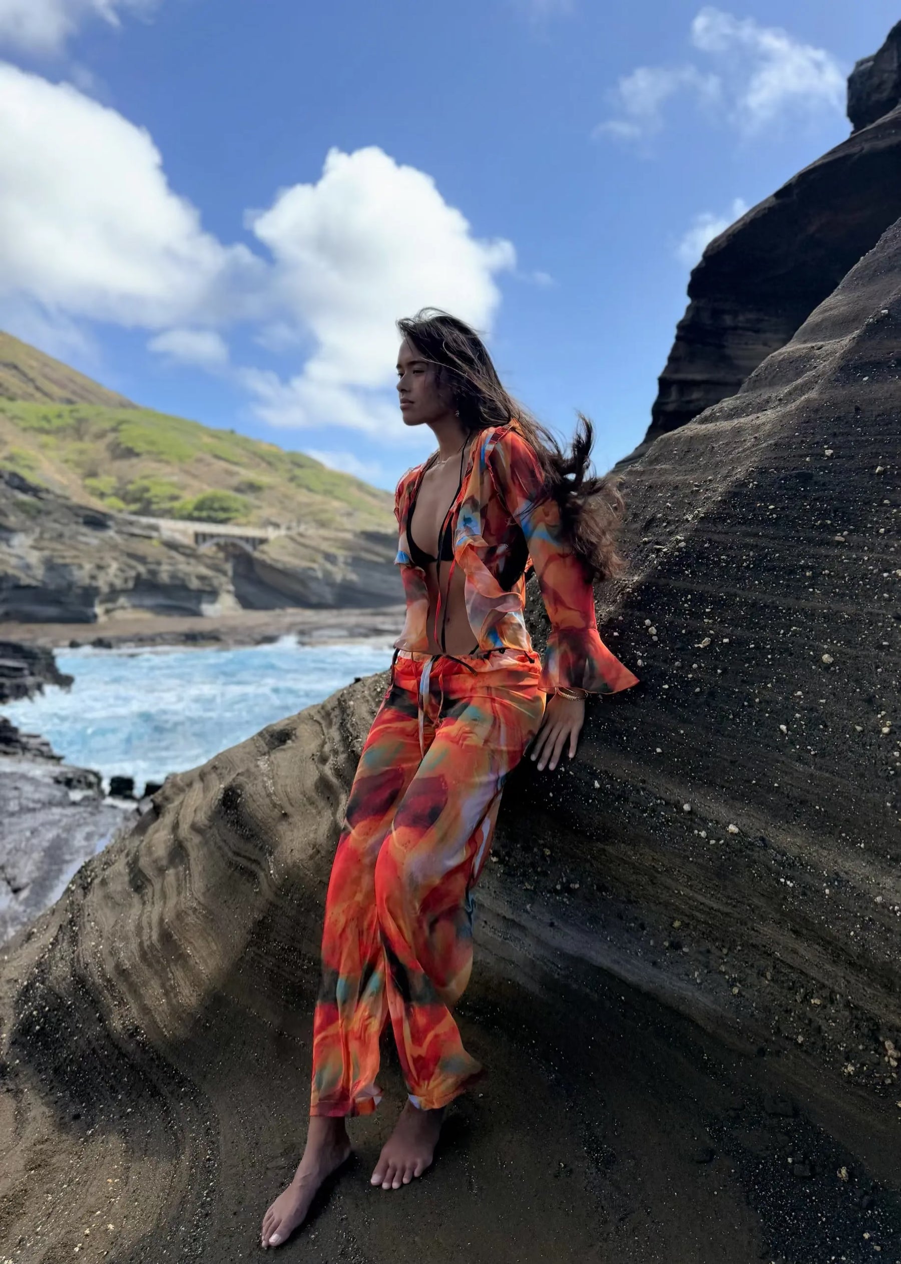 Person in an orange and red outfit sitting on a rocky beach with ocean and sky in the background