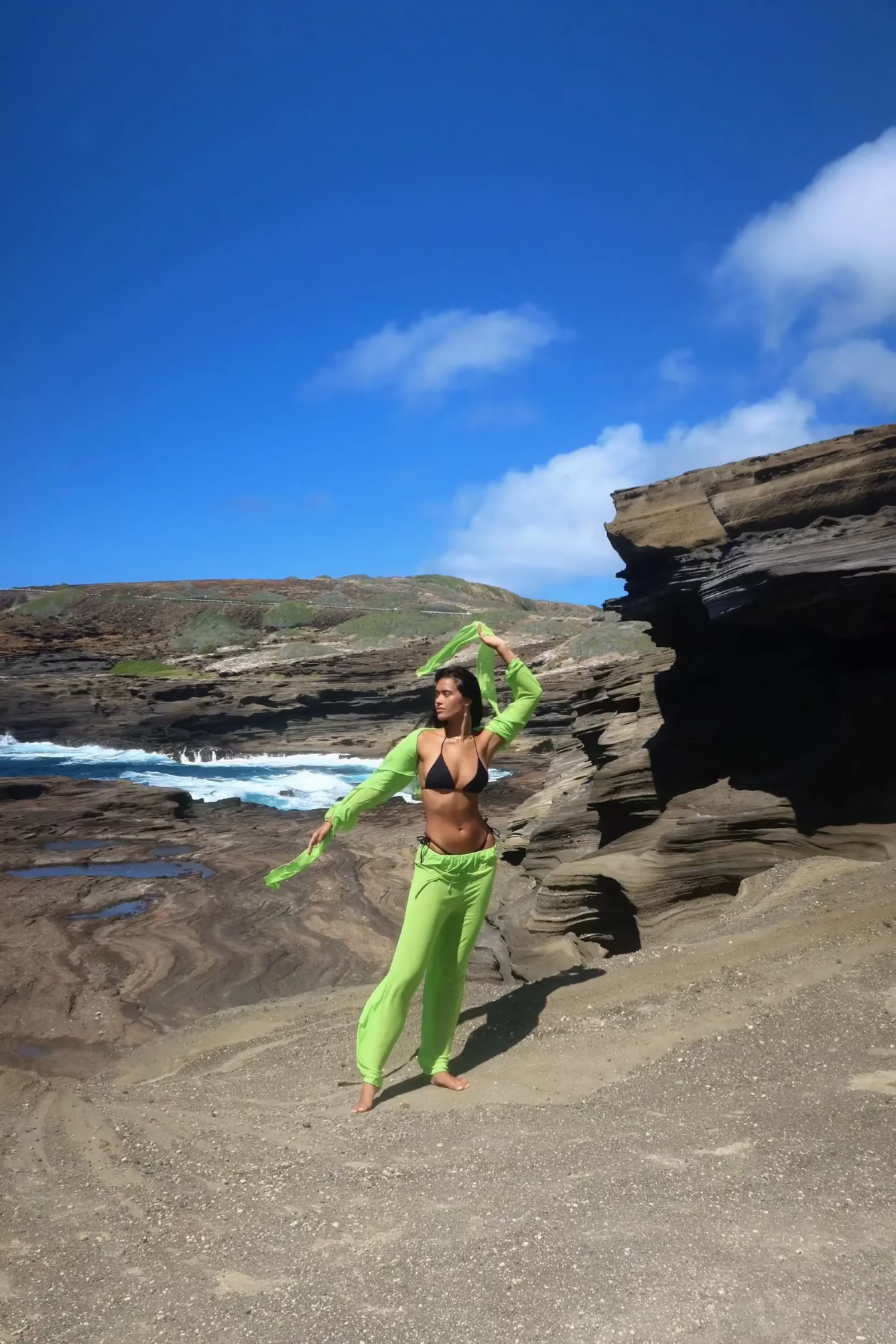 Person in bright green outfit standing on a rocky beach with ocean and cliffs in the background
